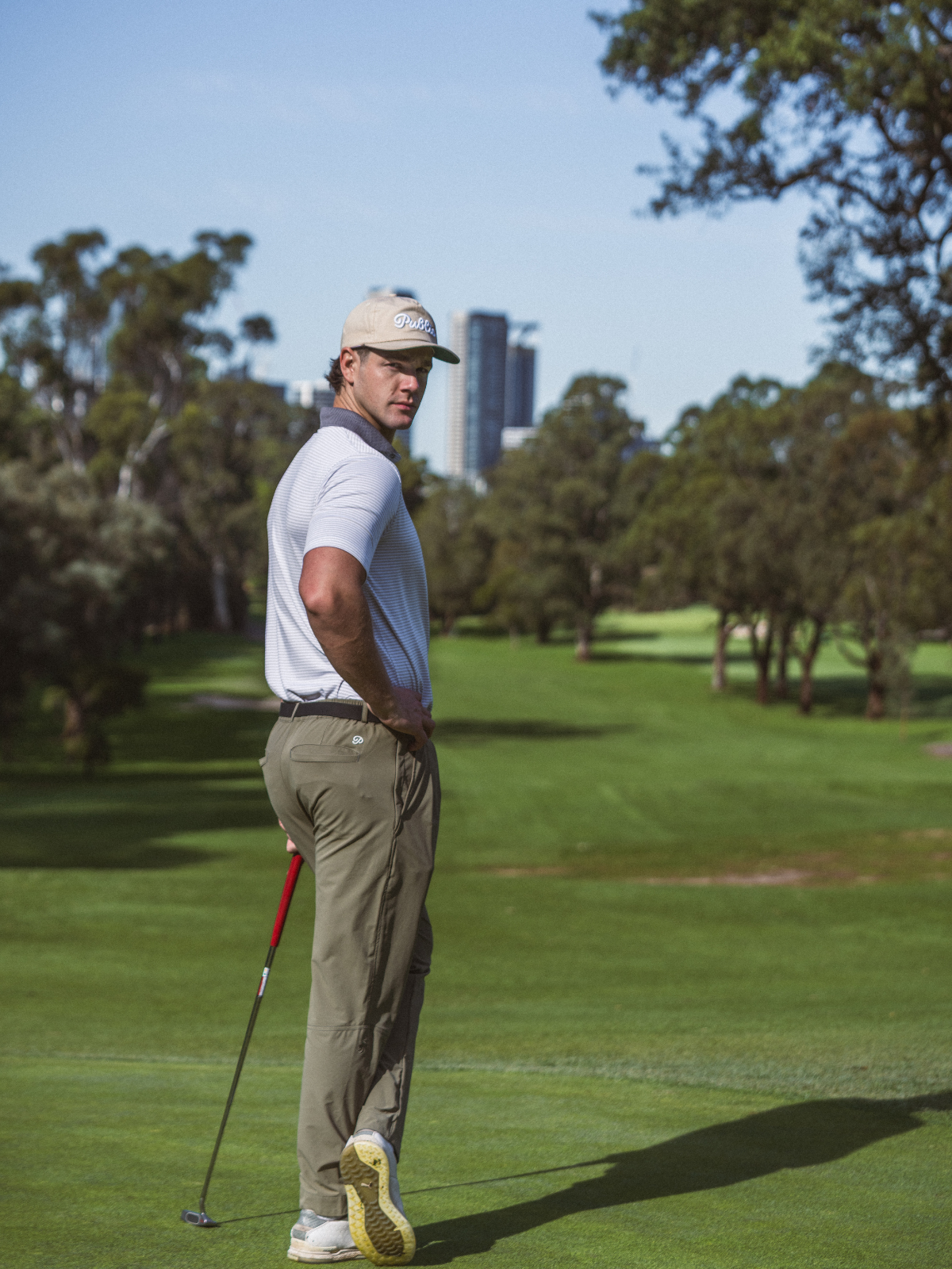 Man wearing Public Drip Anywhere Pleated Pants in olive, three quarter back shot on golf course with city skyline