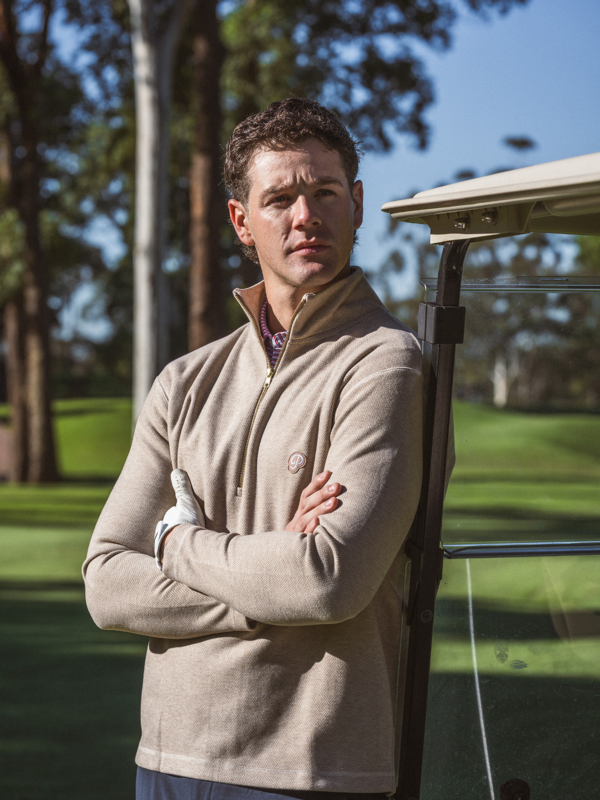 Man wearing Public Drip Herringbone Half Zip in tan, leaning against golf cart on course