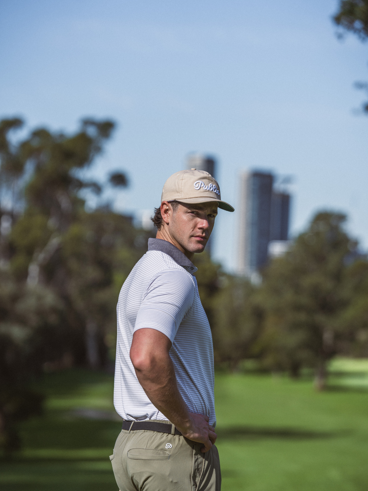 Man wearing Public Drip Public Rope Hat in sand on golf course with city skyline in background
